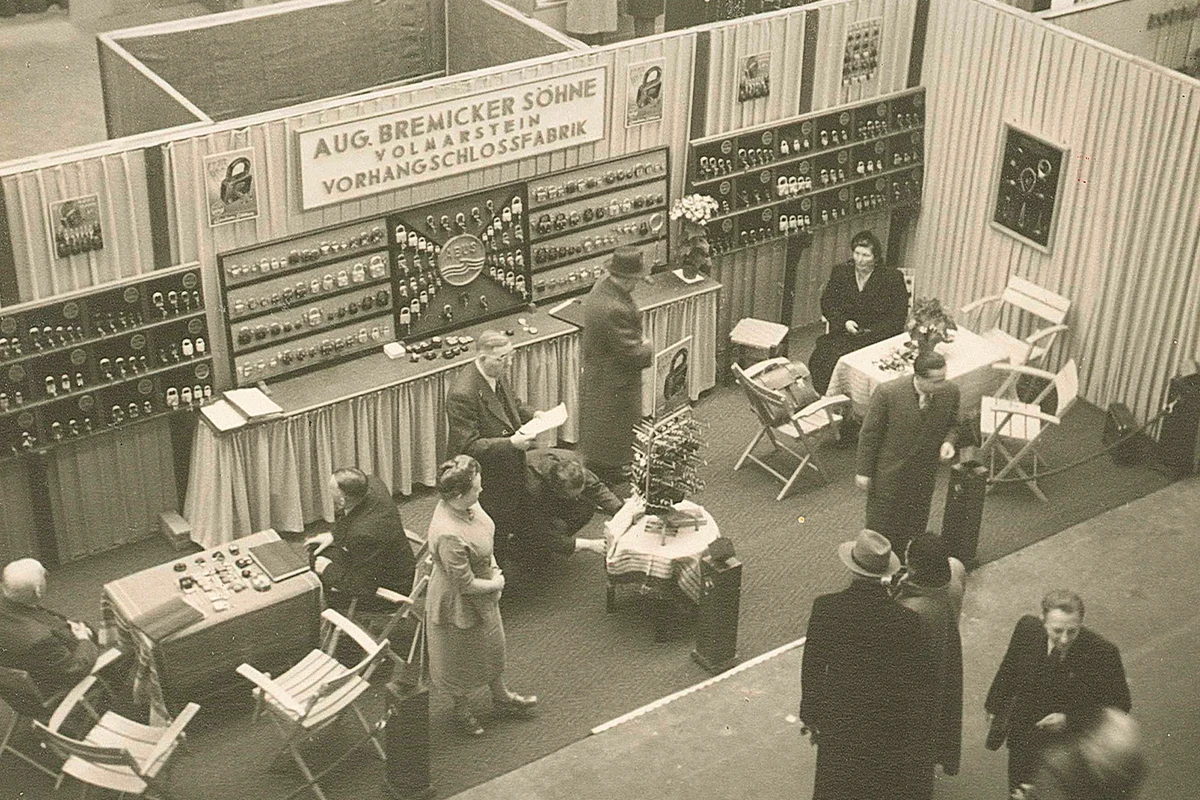 Un stand d'exposition dans les années 1950, avec des cadenas présentés suspendus aux murs et quelques tables sur lesquelles des personnes sont assises ou debout, parfois en train de discuter © ABUS