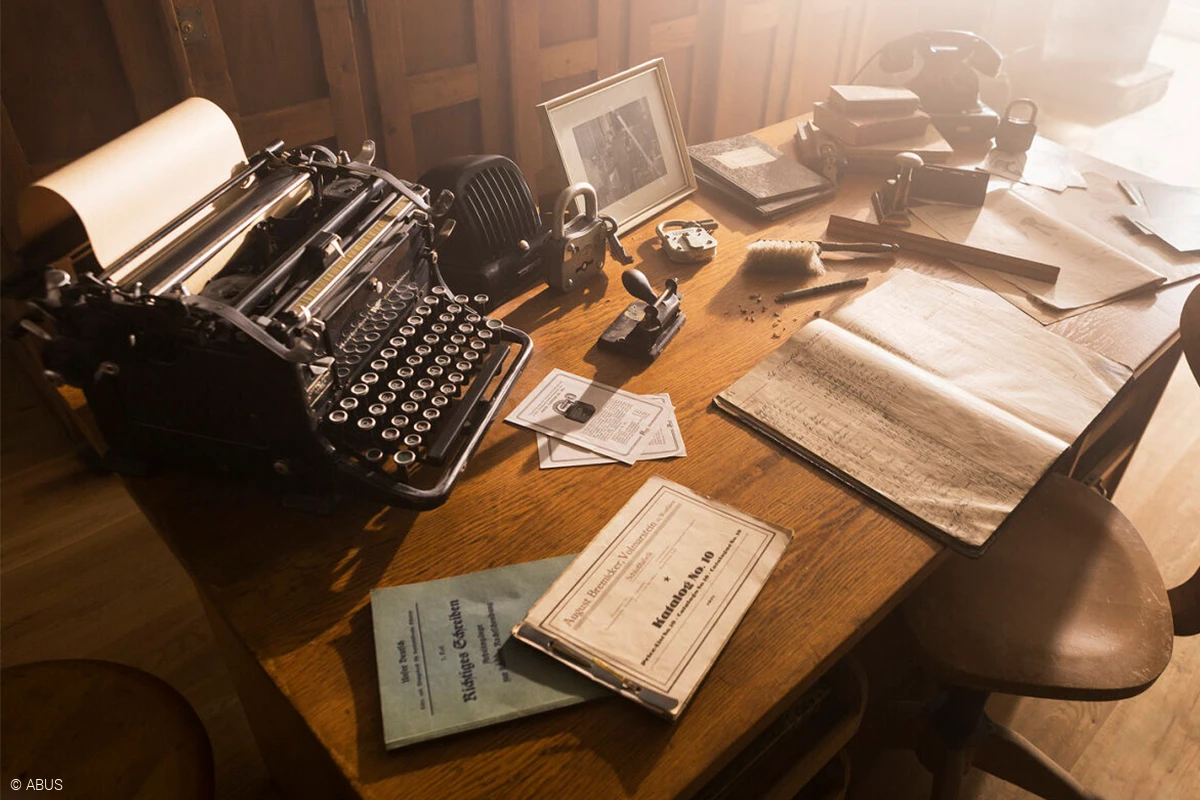 Una scrivania in legno con vari raccoglitori e documenti, una macchina da scrivere, un vecchio telefono, una cornice e alcuni vecchi lucchetti © ABUS