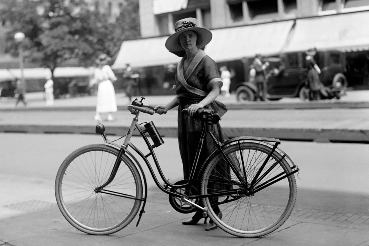 Signora con cappello dietro una bicicletta da donna che guarda direttamente nella macchina fotografica. L'ambiente circostante è sfocato © shutterstock - Everett Collection