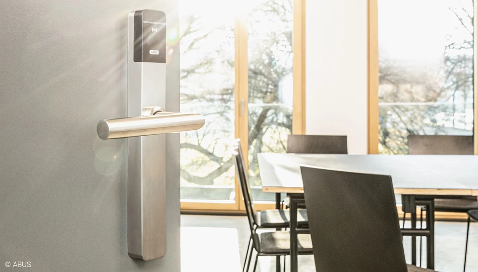 Im linken Bilddrittel eine halbgeöffnete Bürotür mit TECTIQ Beschlag: dahinter ist ein moderner Besprechungsraum mit Tisch und Stühlen sichtbar. © ABUS