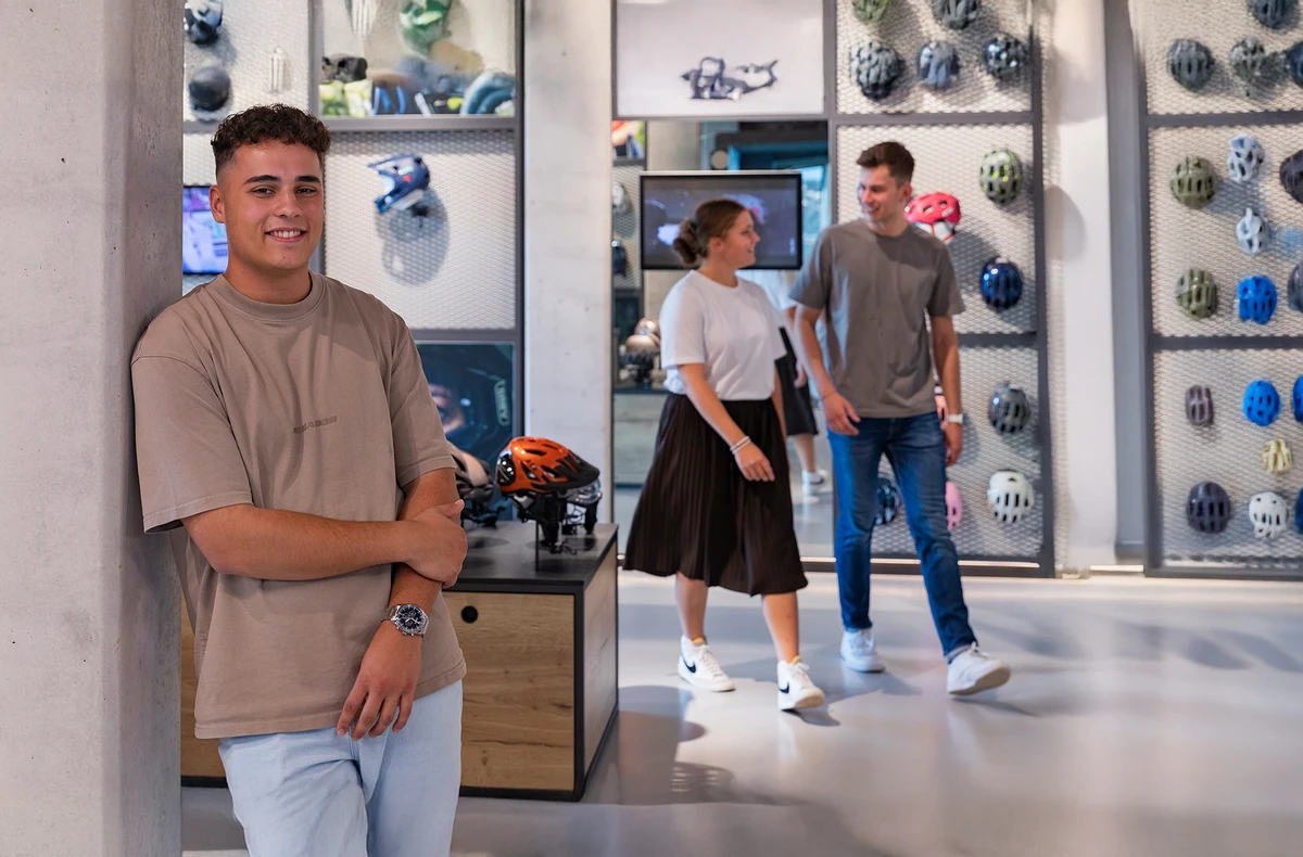 Auszubildender im ABUS Showroom vor einer Helm-Ausstellung, Kolleg:innen im Hintergrund © ABUS