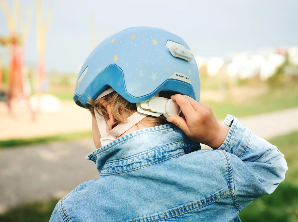 Kind trägt hellblauen Kinderfahrradhelm ABUS Smiley 3.0 FL.ACE LED pure bluestars und justiert Verstellrad am Hinterkopf.