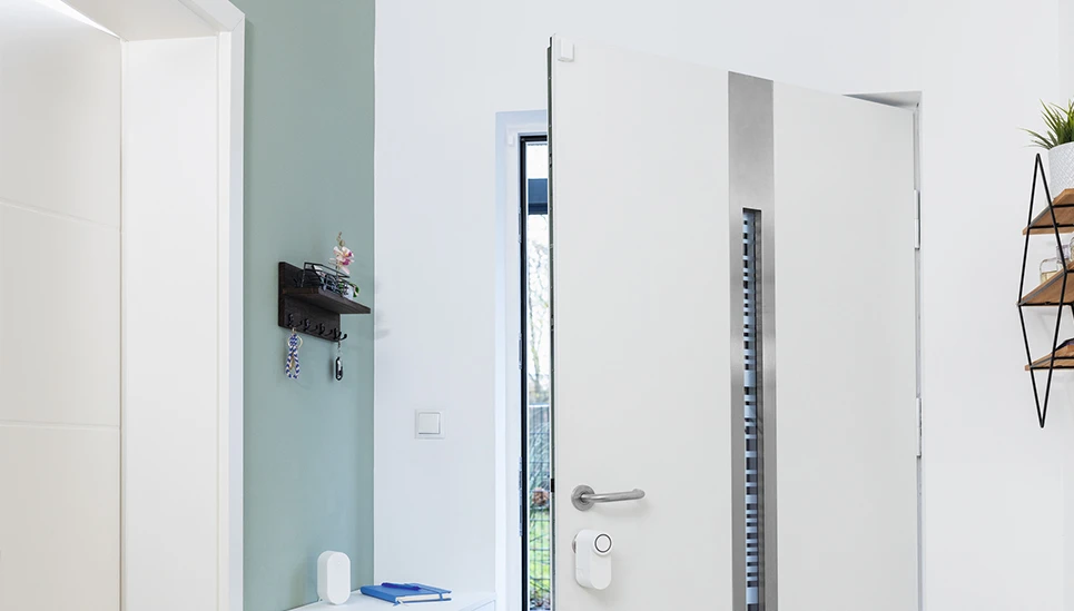 Una porta d'ingresso aperta con i modelli LOXERIS One e SENSOR One installati. Accanto alla porta d'ingresso, un BRIDGE One è appoggiato sopra un armadio © ABUS
