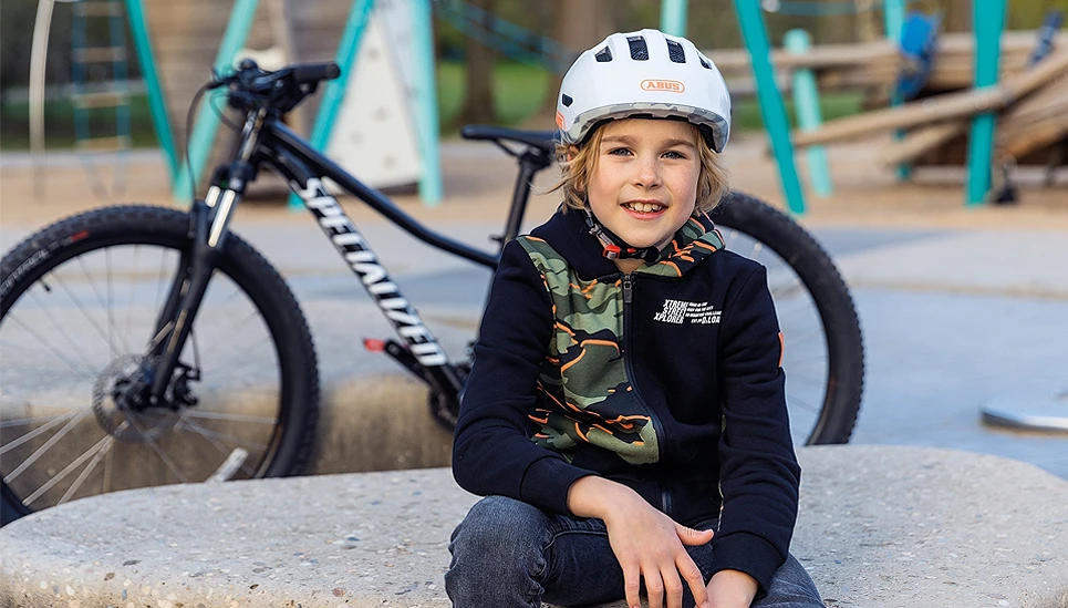 Kind auf dem Spielplatz sitzt mit Fahrradhelm vor einem Fahrrad © ABUS