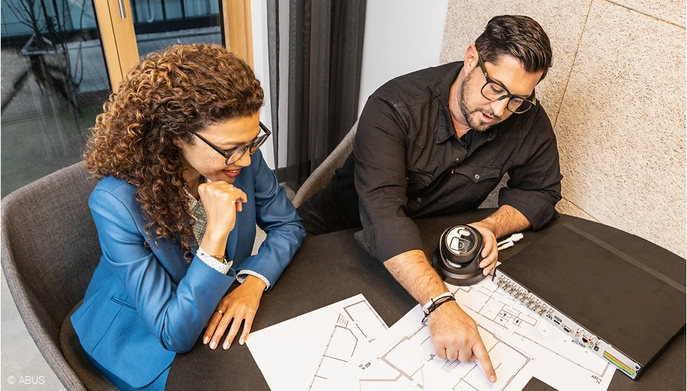 Ein Mann und eine Frau betrachten einen Gebäudeplan. Der Mann (rechts) hält eine Kamera in der Hand und zeigt auf dem Plan den idealen Installationsort dafür.  © ABUS