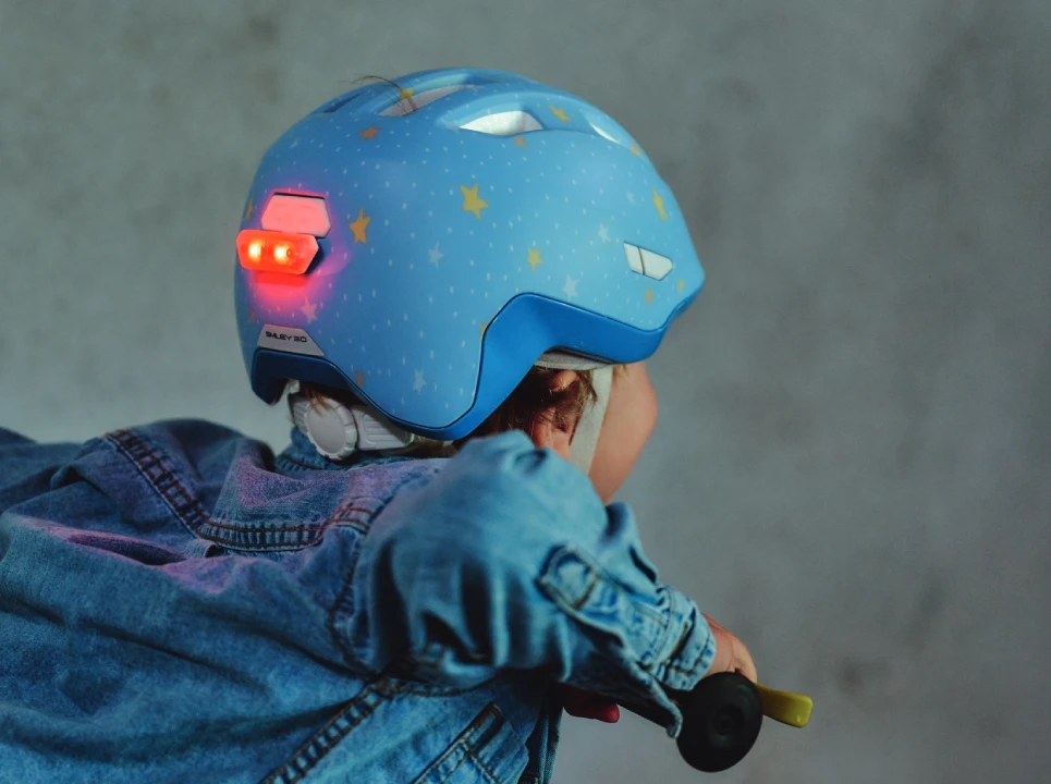 Kind mit ABUS Smiley 3.0 FL.ACE LED Kinderfahrradhelm in der Farbe Pure Bluestars, mit leuchtendem LED-Rücklicht am Hinterkopf © ABUS