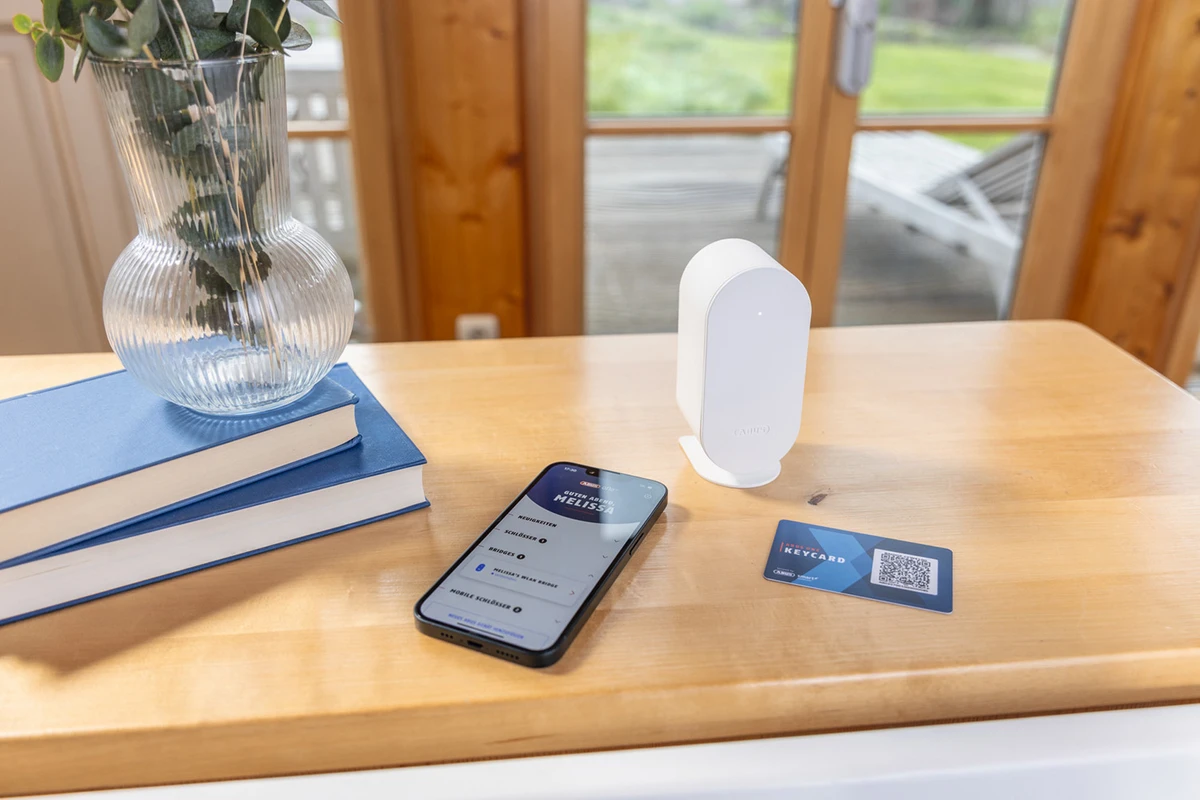La BRIDGE One est posée sur une table, à côté d'un smartphone avec l'application ABUS One ouverte et d'une carte-clé © ABUS