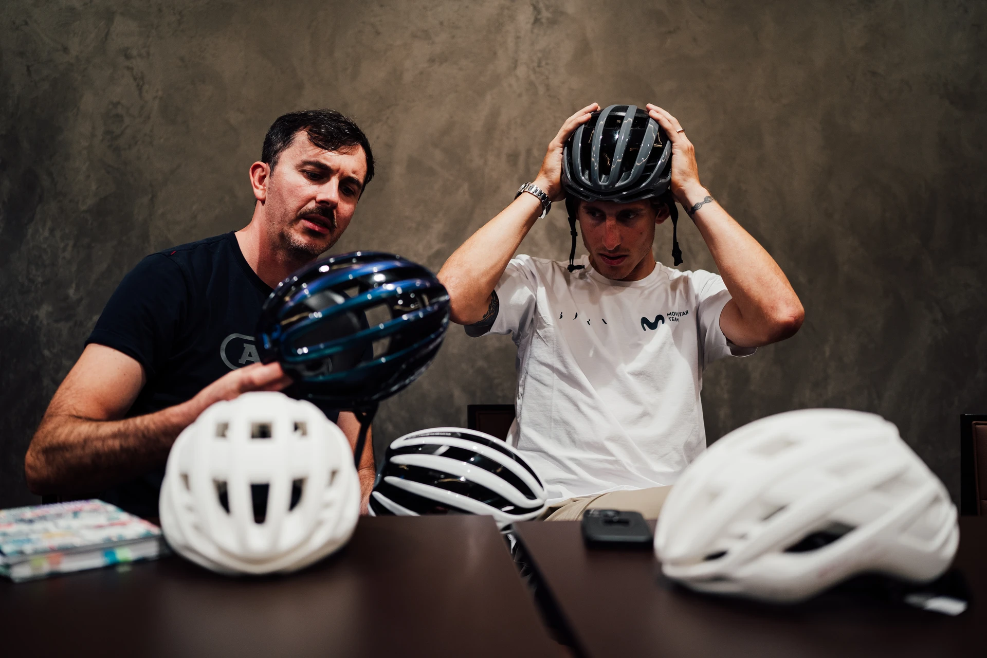 Product manager and a professional cyclist from the Movistar team sit at the table and scrutinise several AirBreaker 2.0 racing helmets © ABUS