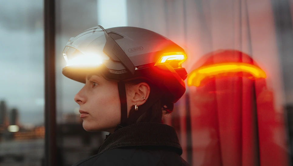 Urban Fahrradhelm HYP-E mit Visier und integriertem Front- und Rücklicht; urbane Radfahrerin im Profil bei Dämmerung vor Glasfassade © ABUS