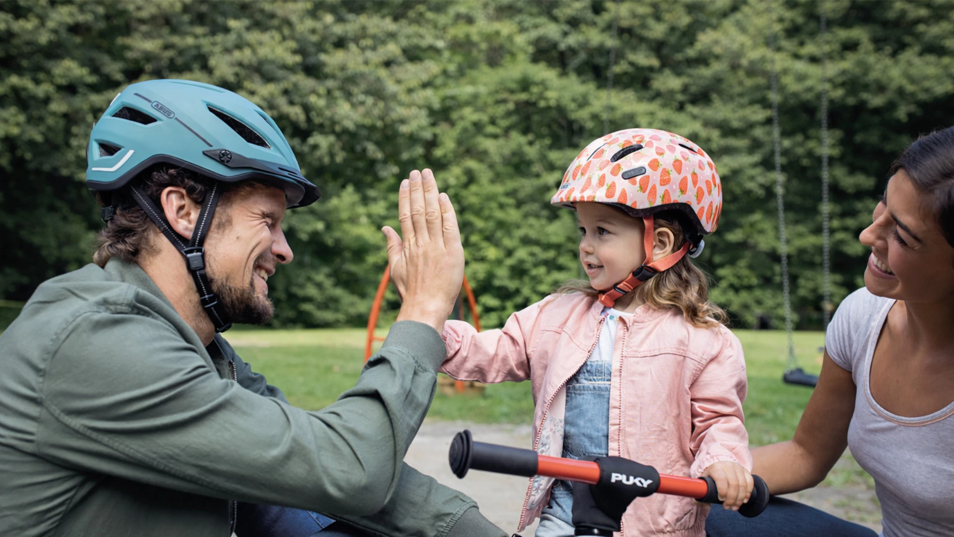 Gemeinsamer Moment im Park: Ein Kind auf dem Laufrad wird von Erwachsenen unterstützt und angelächelt © ABUS