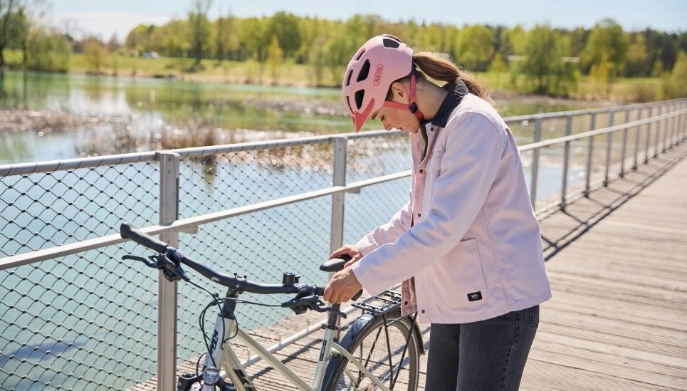 A young woman is standing next to her bike by a lake, looking down. She is wearing a YOUN-I 3.0 ACE bike helmet in powder coral © ABUS