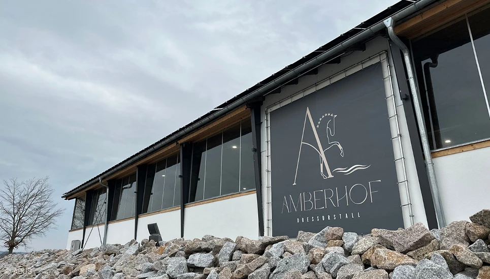 Zweigeteiltes Bild: im linken Bilddrittel das Logo des Dressurstalls Amberhof, im rechten Bildteil Blick in die Stallgasse. © ABUS