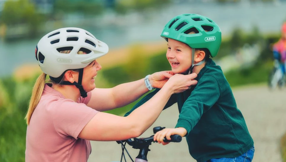 A mother and son smile at each other as she fastens his cycling helmet. Both are wearing the YOUN-I 3.0 cycling helmet © ABUS