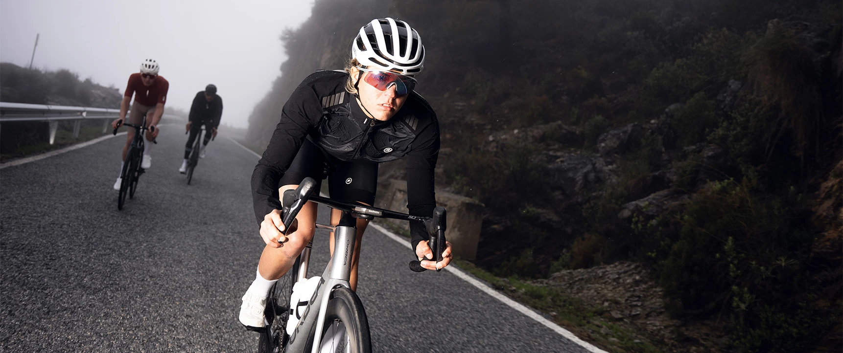 Radfahrerin mit dem Rennradhelm AirBreaker 2.0 in Shiny White in Aero-Position auf nebeliger Bergstraße, zwei weitere Radfahrer im Hintergrund © ABUS