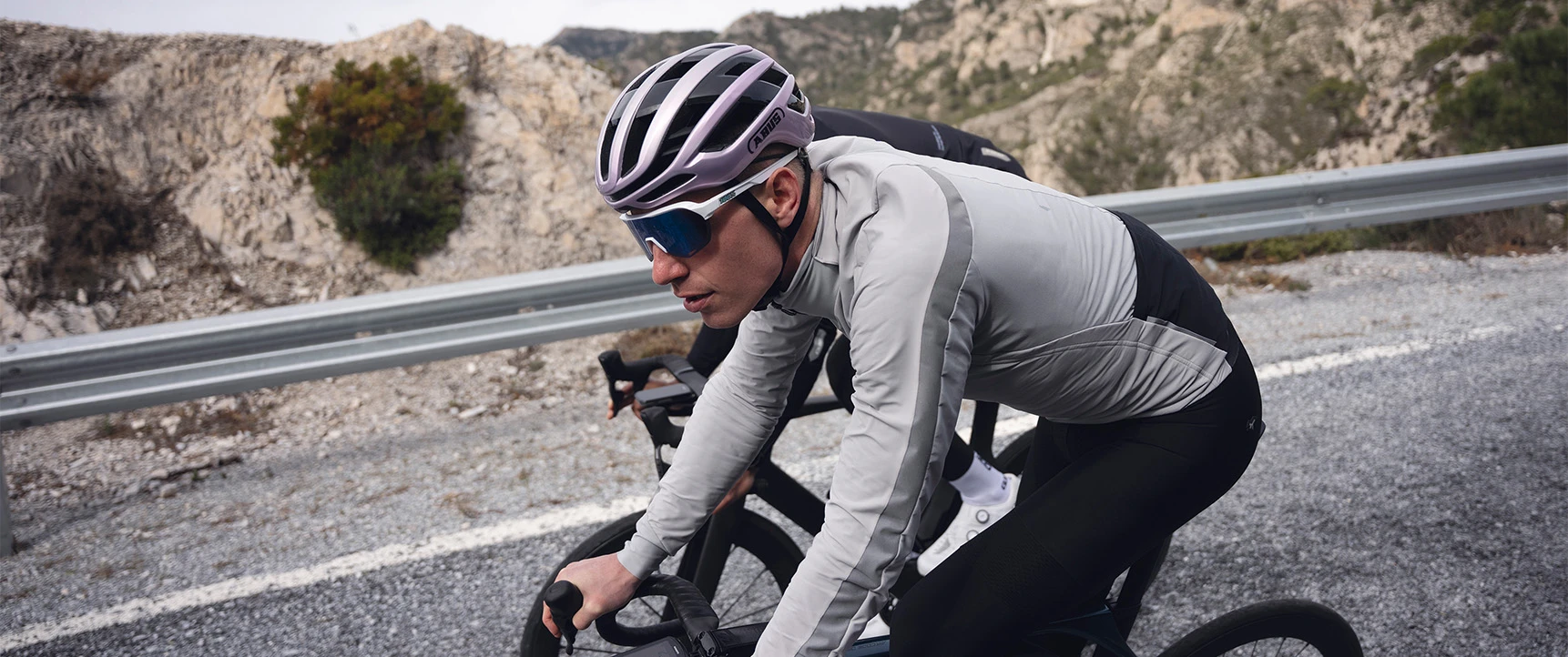 Road cyclist with the AirBreaker 2.0 road helmet in purple riding on a winding mountain road, rocky landscape and guardrail in the background © ABUS