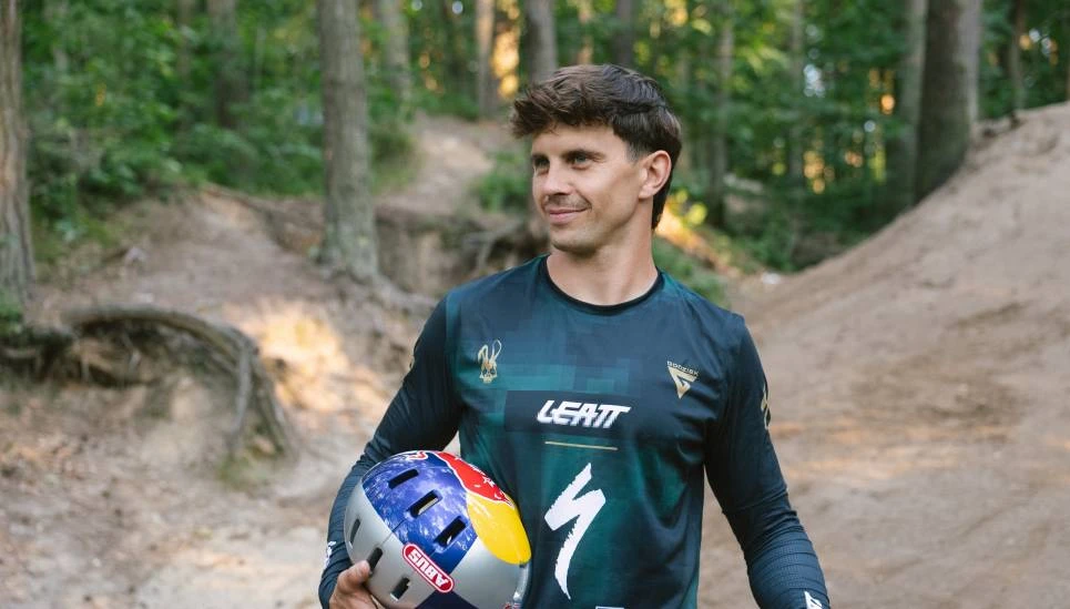 Szymon Godziek trzyma w ręku kask rowerowy i spogląda w dal na leśnej ścieżce. © ABUS