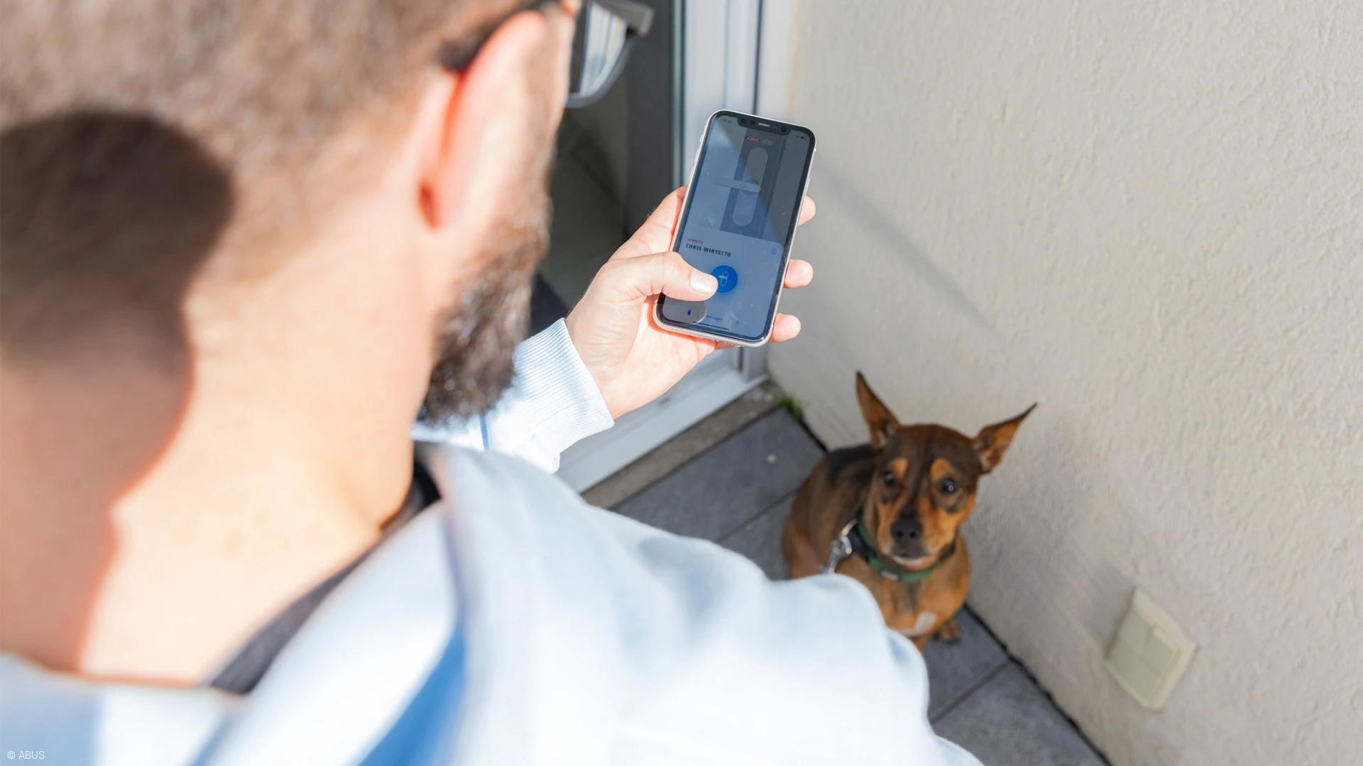 Mann öffnet mit ABUS One App seine Terrassentür während sein Hund ihn erwartungsvoll anschaut © ABUS