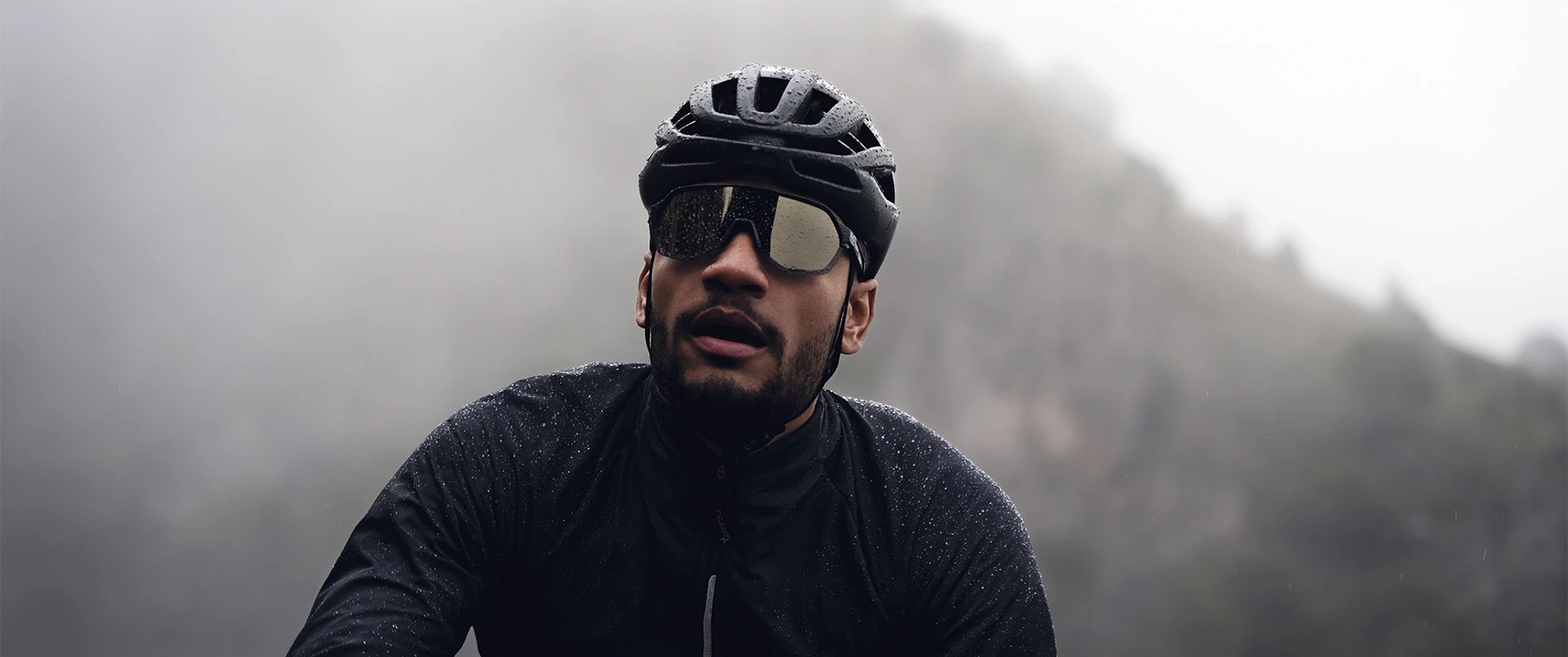 Cyclist with the AirBreaker 2.0 road helmet in Velvet Black riding in the rain on a foggy mountain road, close-up with reflective glasses © ABUS