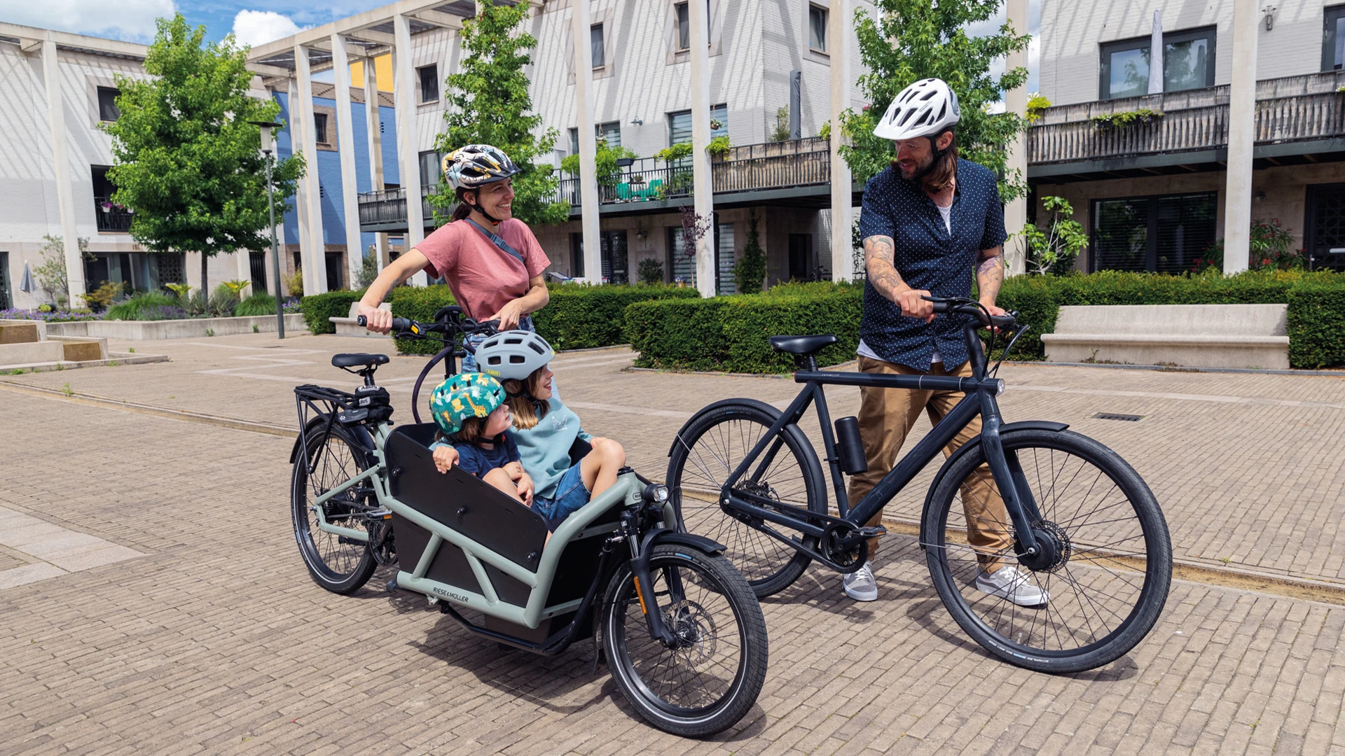 Zwei Erwachsene mit Fahrrädern auf einem urbanen Platz; Frau fährt ein Lastenrad mit zwei Kindern, alle tragen Fahrradhelme von ABUS © ABUS