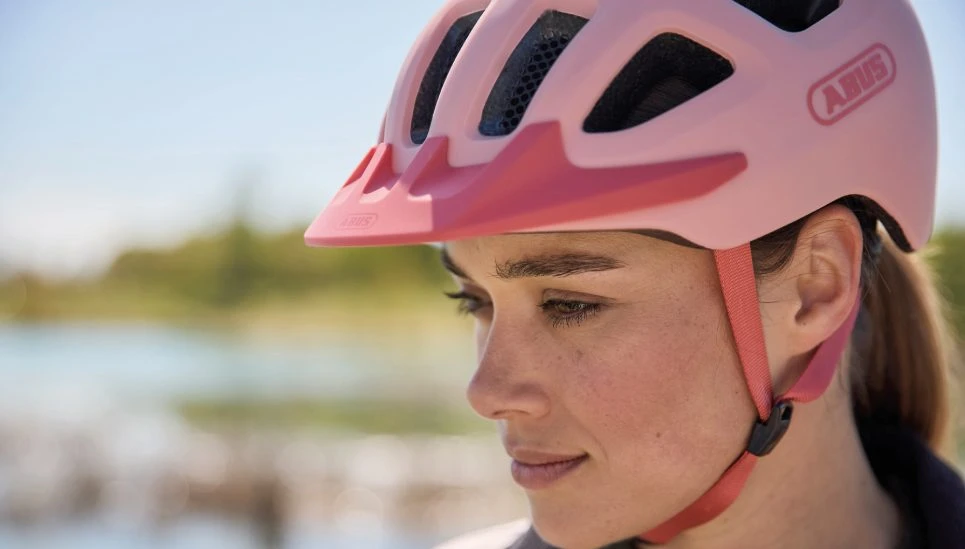 Close-up of a woman wearing a YOUN-I 3.0 “powder coral” bicycle helmet with the chin strap fastened. She is looking down; in the background is a blurred landscape with water © ABUS