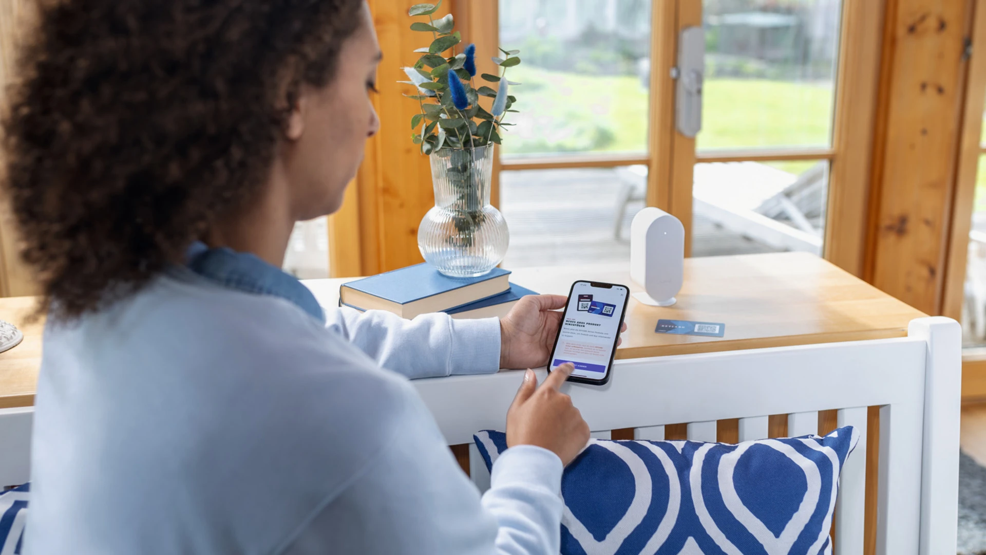 A woman holds a smartphone with the ABUS One app open in her hand, while a keycard and a Bridge One sit on the sideboard in front of her © ABUS