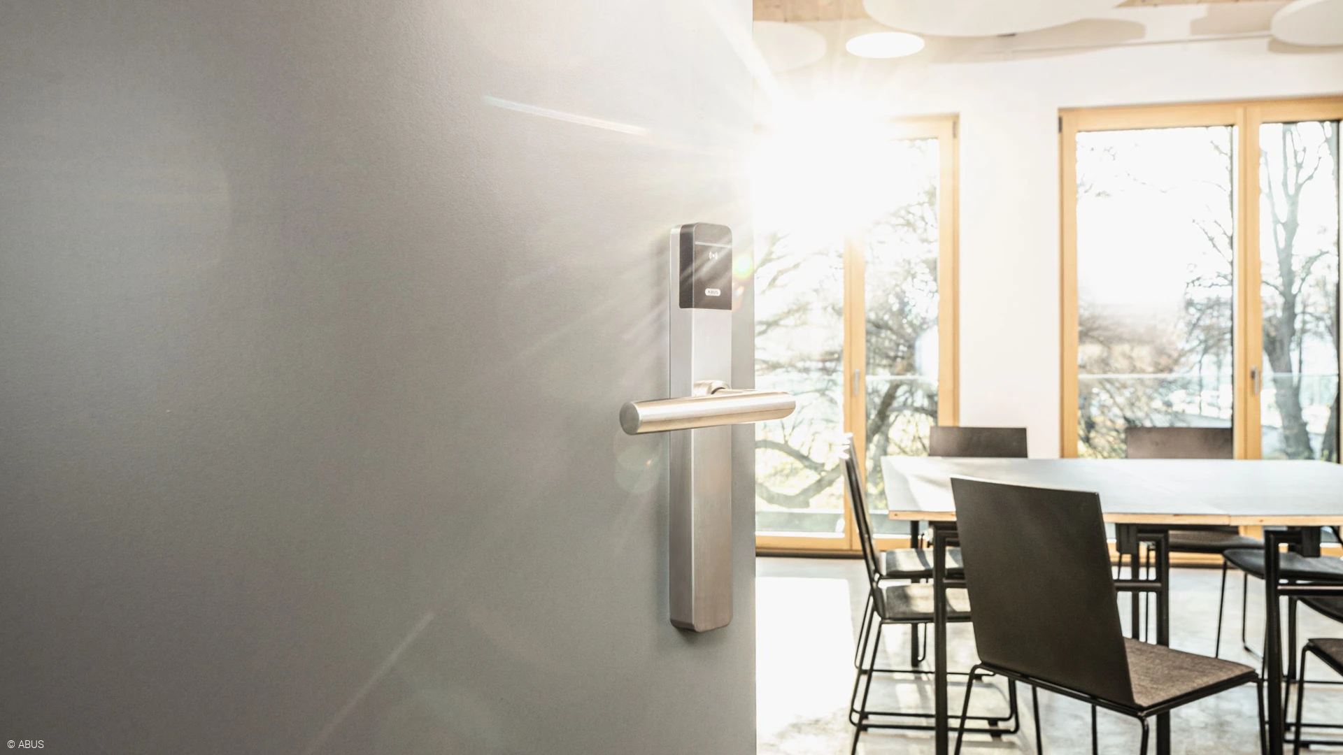 Dans la moitié gauche de l'image, une porte de bureau moderne entrouverte avec une ferrure TECTIQ. A droite, une table et des chaises de bureau.