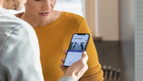 Ein Mann hält sein Smartphone in der Hand. Auf dem Display sieht man die Livebilder seiner ABUS Akku Kamera Pro. Eine Frau, die neben ihm steht, blickt ebenfalls auf das Display.