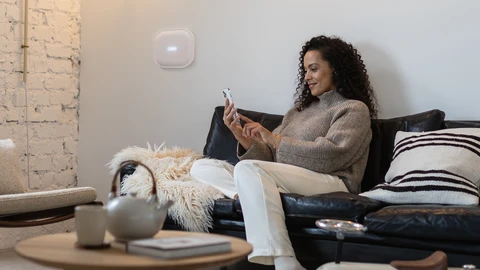 Frau auf Sofa sitzend betrachtet entspannt lächelnd ihr Smartphone. Auf der Zimmerwand im Hintergrund ist die Comfion Pro-Zentrale montiert.