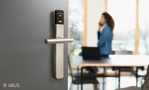 En primer plano, una puerta de oficina moderna con herrajes TECTIQ. Al fondo, una mujer hablando por teléfono.