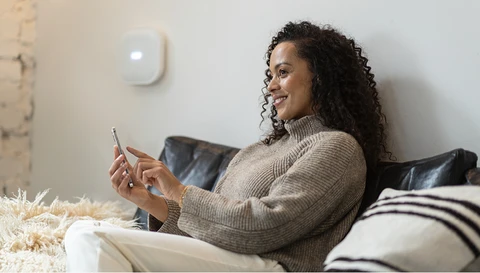 Frau sitzt entspannt lächelnd auf einem Sofa, ein Handy in der Hand. An der Wand im Hintergrund ist die Comfion Pro Zentrale zu sehen.