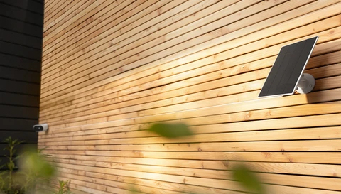 Am rechten Bildrand sieht man das Solarpanel, das durch ein Kabel mit der Akku Kamera Pro am linken Bildrand verbunden ist. Beide Produkte sind an einer Holzwand montiert.