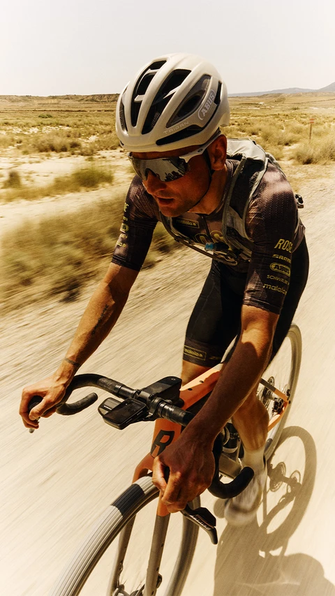 Cycliste professionnel sur une piste naturelle poussiéreuse dans un paysage vallonné avec le casque de vélo de route Taipan © ABUS