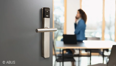 A kép bal oldalán egy félig nyitott, modern irodai ajtó látható, TECTIQ-zárral. A kép jobb oldalán egy asztal és irodai székek láthatók.
