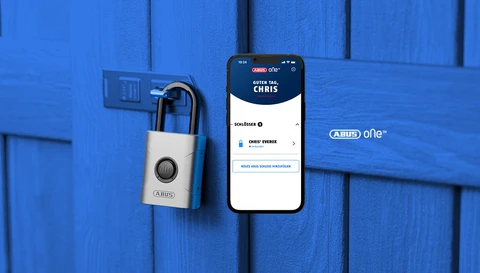 EVEROX One intelligens lakat egy fából készült fészer ajtaján és egy okostelefon az alkalmazás képernyőjével az előtérben © ABUS