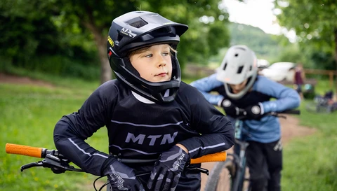Kind im Fullface-MTB-Helm ABUS YouDropp FF in Velvet Black, stützt sich auf den Lenker; weiteres Kind mit Helm im Hintergrund © ABUS