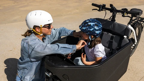 Woman wearing the urban bicycle helmet XOXO while adjusting the Smiley 3.0 blue whale children's helmet for her child in a cargo bike. © ABUS