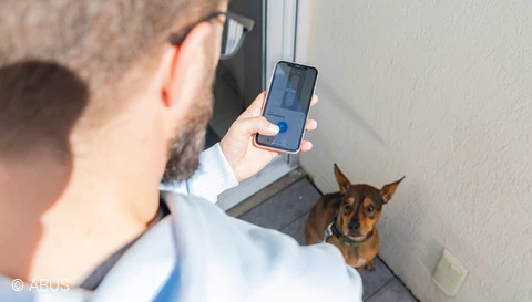 Mann öffnet mit ABUS One App seine Terrassentür während sein Hund ihn erwartungsvoll anschaut © ABUS