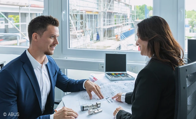 Ein Mann (links) plant mit einer Frau (rechts) anhand des Bauplans die Zutrittskontrolle für einen Neubau. Die Baustelle ist durch das Fenster zu sehen. © ABUS