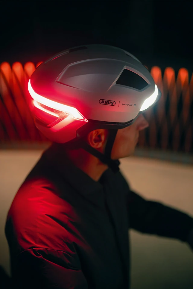 Eine Person trägt den HYP-E Fahrradhelm in der Farbe gleam silver mit leuchtendem roten Rücklicht. Der Hintergrund zeigt unscharfe, orangefarbene Lichter © ABUS