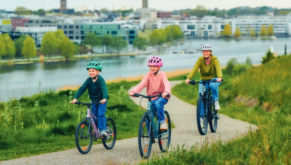 A woman with two children is cycling along the lake. They are all wearing the YOUN-I 3.0 cycle helmet in different colours © ABUS