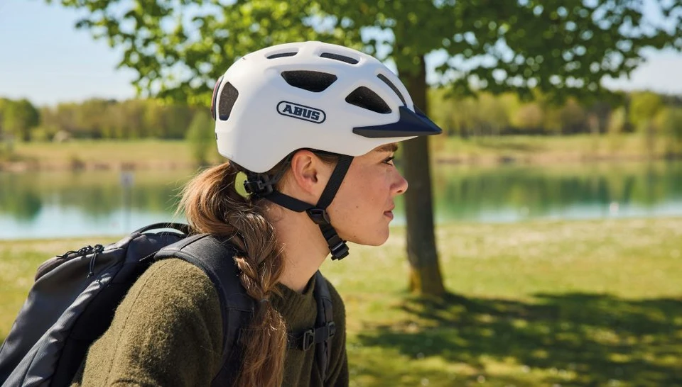 Side view of a woman wearing a YOUN_I 3.0 ACE bicycle helmet in off-white. She is carrying a black backpack and riding against a green landscape © ABUS
