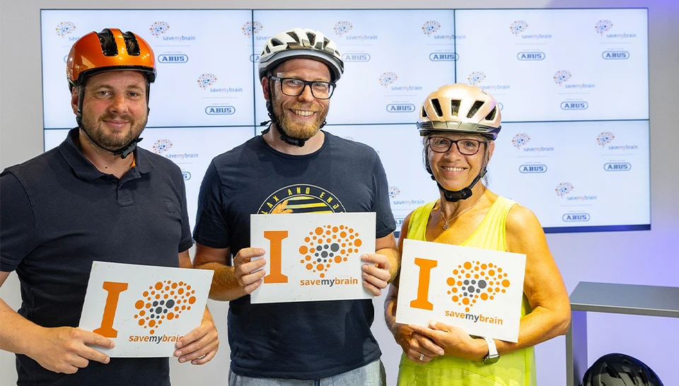 Drei ABUS Mitarbeiter nebeneinander mit Fahrradhelm und einem Schild mit claim "I savemybrain" &copy; ABUS