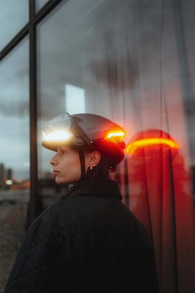 Side view of a woman wearing the HYP-E bike helmet in Volcano Titan, with integrated visor and glowing red rear light, standing in front of a glass wall © ABUS