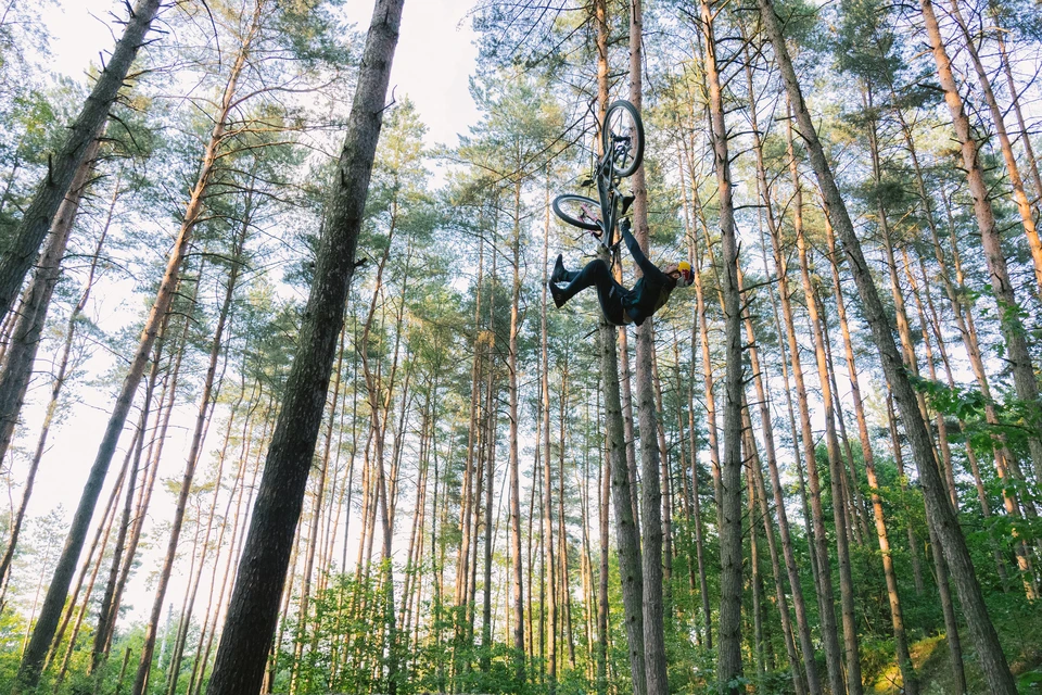 Szymon Godziek wykonuje spektakularny backflip na rowerze górskim wysoko nad leśną trasą © ABUS