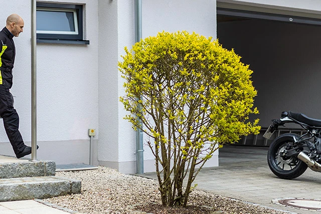 Ein Mann in einem schwarzen Motorrad-Overall verlässt sein Haus und nähert sich seinem Motorrad, das vor der geöffneten Garage steht. ©ABUS