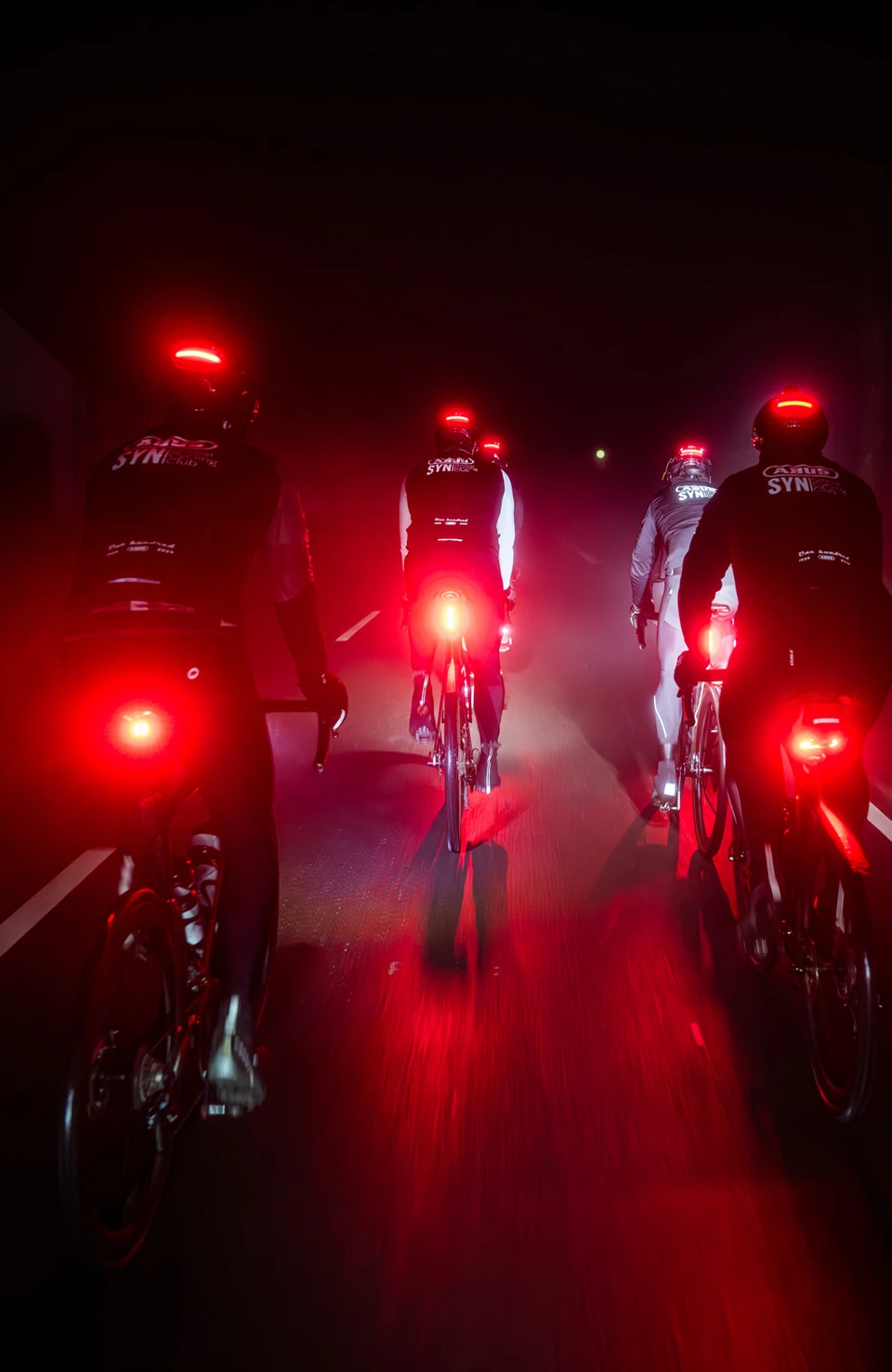 Flere racercyklister med Taipan LED-cykelhjelme kører om natten på vejen, røde baglygter lyser i tågen. © ABUS
