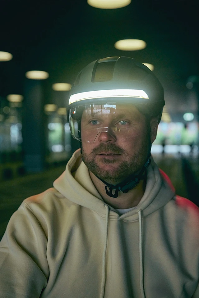Ein Mann trägt den HYP-E ACE Fahrradhelm mit integriertem Visier und reflektierendem LED-Frontlicht. Der Helm leuchtet in einem dunklen, modernen Raum © ABUS