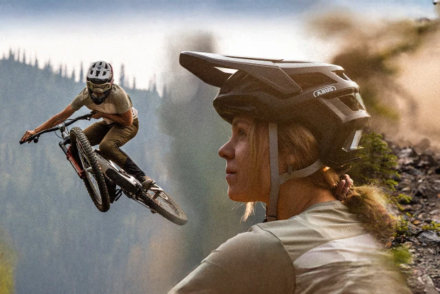 A female mountain biker wearing a TARGON MTB helmet on dusty terrain, with another mountain biker riding through the background in an action-packed scene © ABUS
