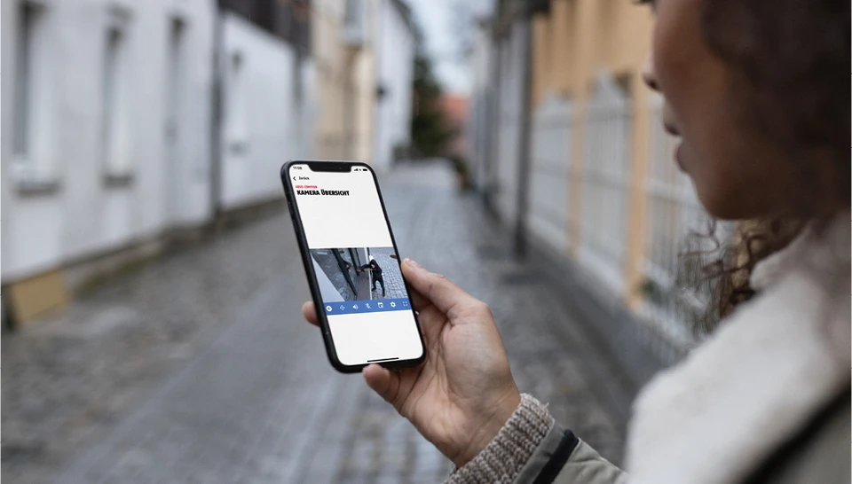 Eine Frau betrachtet unterwegs auf ihrem Smartphone ein Live-Bild von ihrem Zuhause. Ein Alarm wurde ausgelöst, weil sich jemand unbefugt Zutritt verschaffen will.