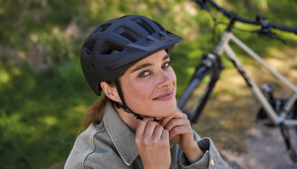 A woman wearing a YOUN-I 3.0 ACE bicycle helmet in black looks at the camera as she fastens the helmet © ABUS